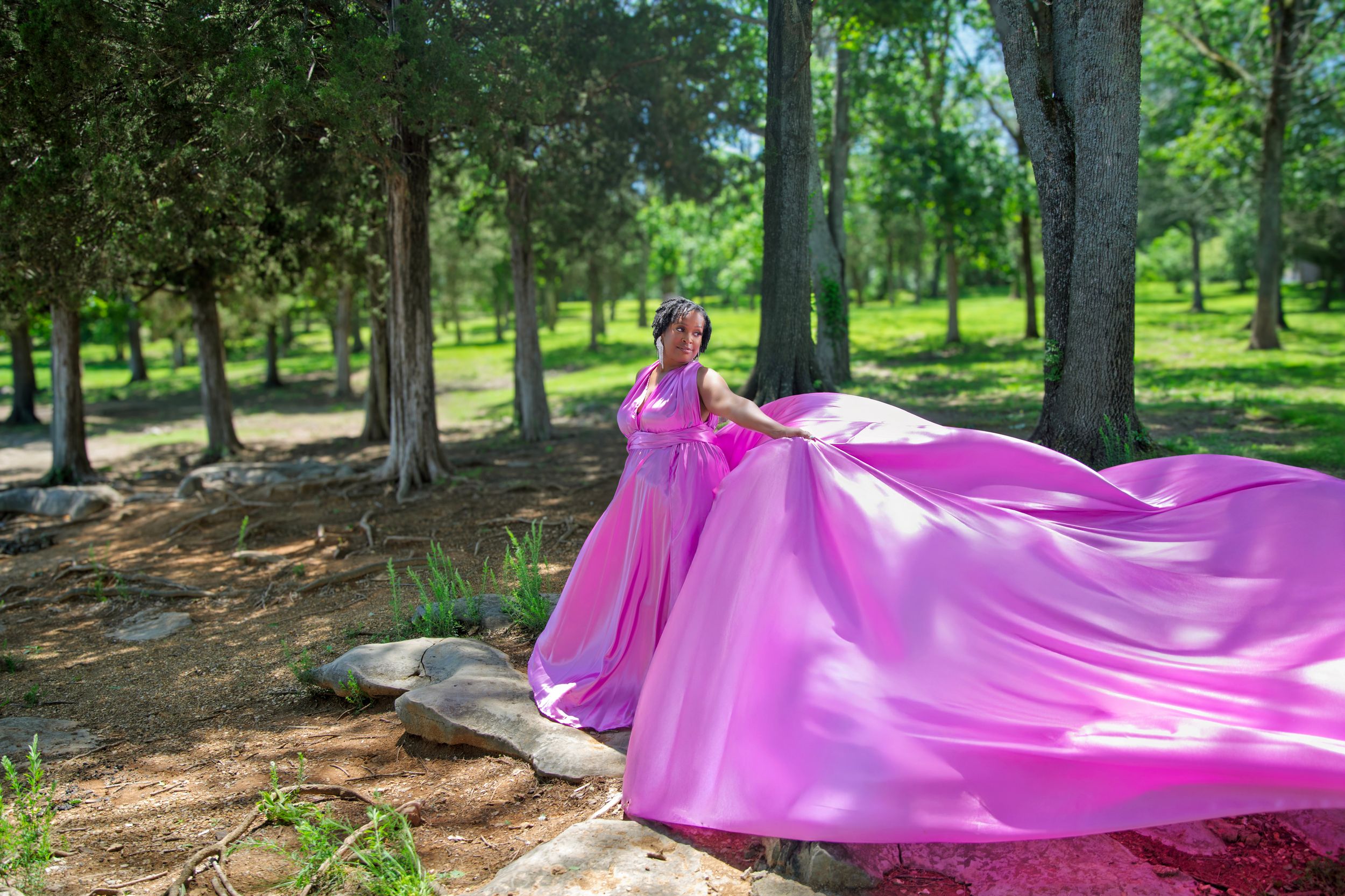 A bright magenta fabric billows dramatically in a green park setting with tall trees casting shadows on the ground.