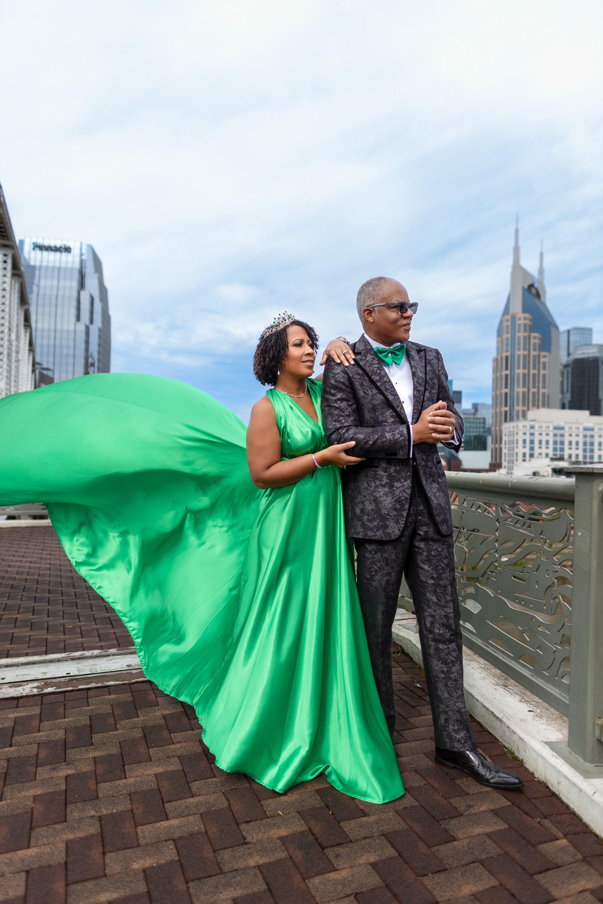 Elegant couple poses on Nashville rooftop with cityscape background, her emerald green gown flowing in the wind.