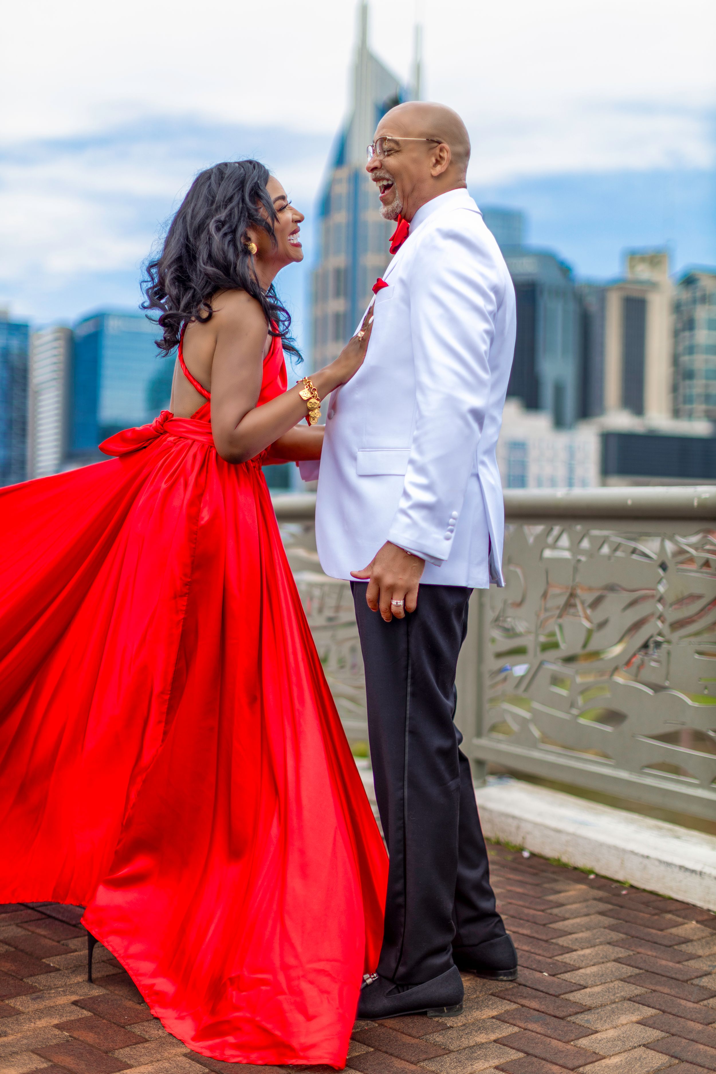 A couple shares a romantic moment on a rooftop terrace, with her in a flowing red gown and him in a white suit jacket.