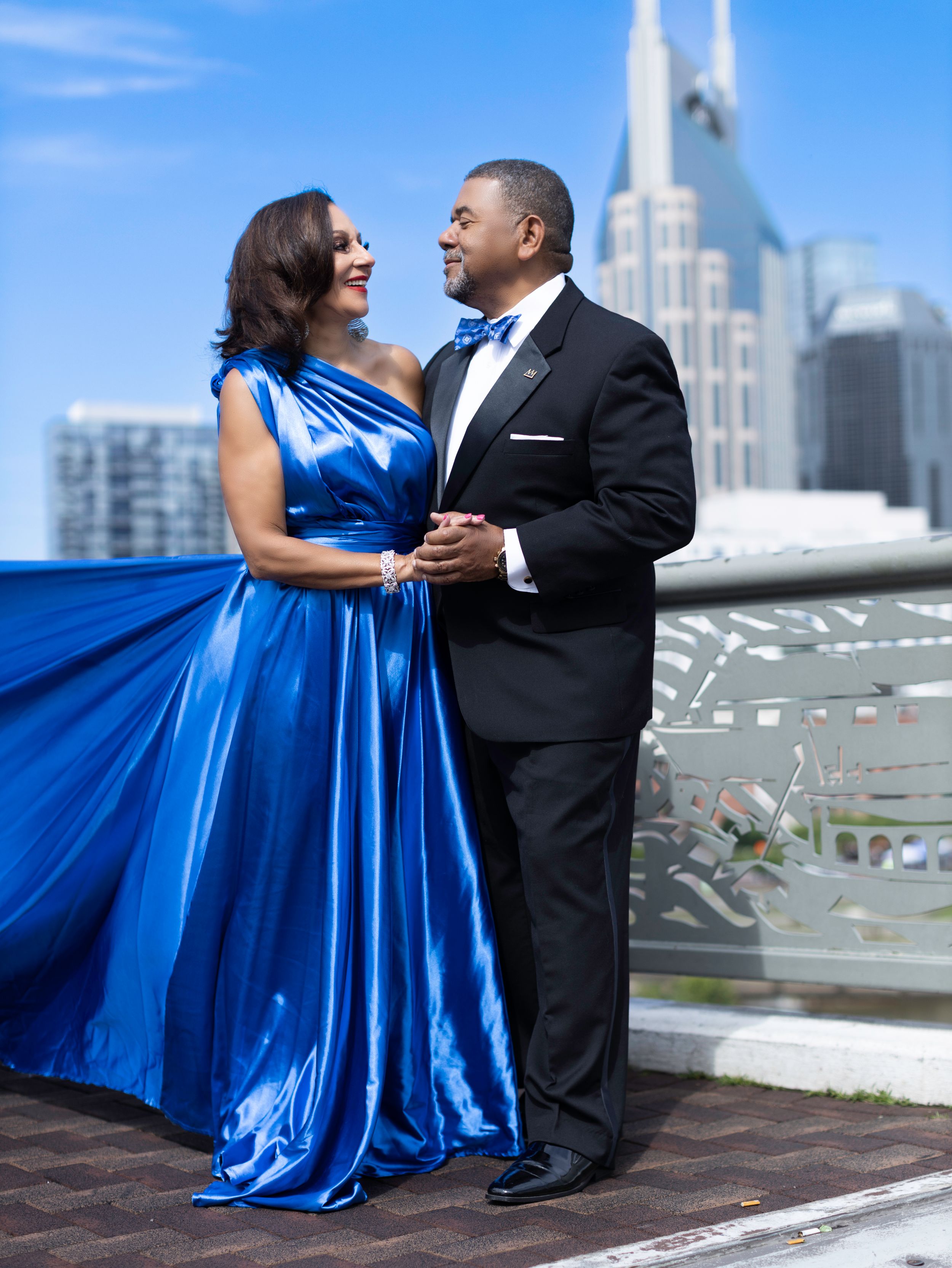 A couple shares a romantic moment in formal attire against a city skyline backdrop, with a flowing blue satin gown catching the light.