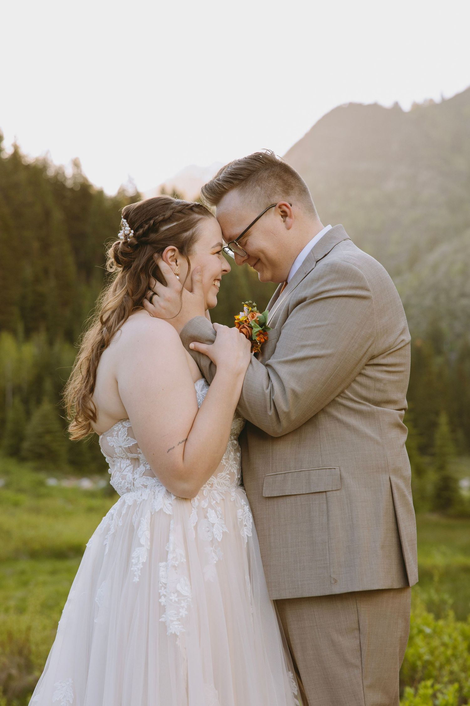 Bride in flowing white dress holds bouquet while standing on wooden deck surrounded by evergreen mountains at sunset.