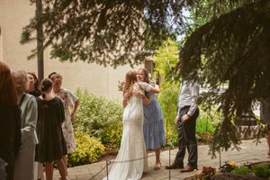 Wedding guests gather under tall trees at an outdoor celebration on a sunny summer day.
