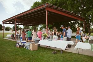 Outdoor gathering under a covered pavilion with people around white tables and boxes during a daytime event.