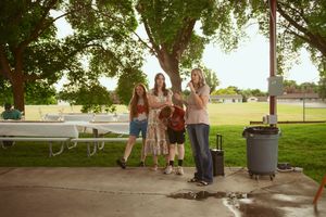 A group of people enjoy a sunny day gathering at a park with picnic tables and trees.