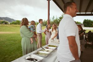 A line of wedding guests gather at a buffet table under a rustic covered pavilion.