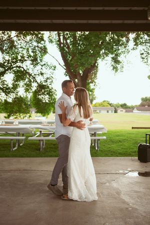 A couple shares a romantic dance moment under a shaded pavilion in a sunlit park with picnic tables in the background.