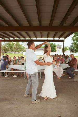 A couple dances romantically under a covered pavilion area while guests watch from picnic tables in the background.