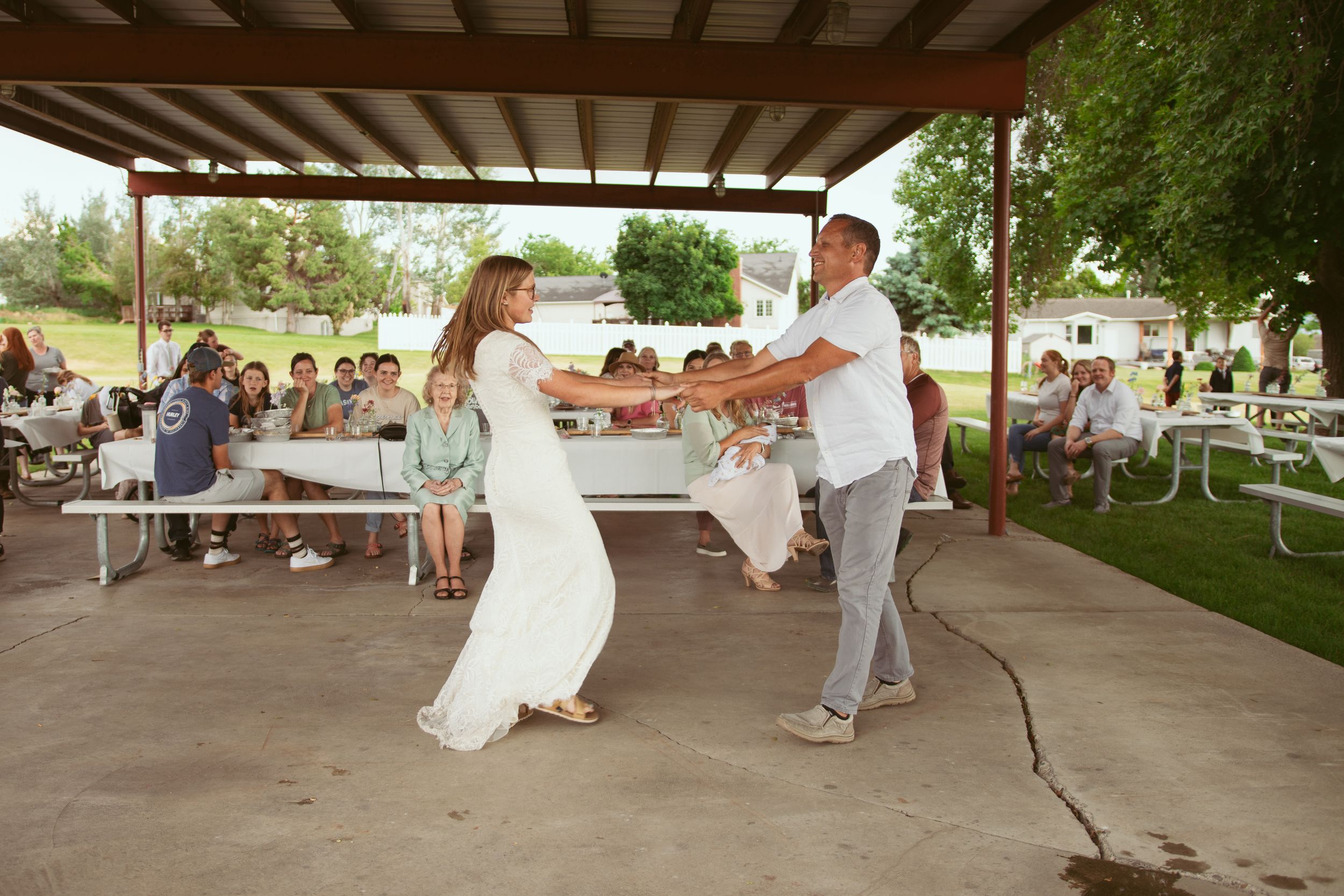 A couple dances together under a covered outdoor pavilion during a celebration with guests seated in the background.