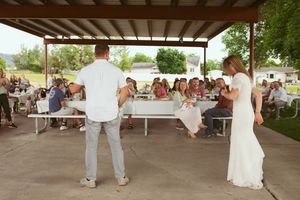 Casual outdoor wedding reception under a covered pavilion with guests seated at long tables in a park setting.