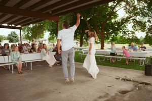A couple performs their first dance under a covered outdoor space while guests watch from picnic tables.