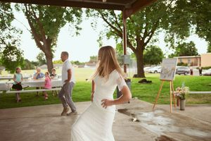 A couple dances together under a shaded pavilion at an outdoor event with trees and people in the background.