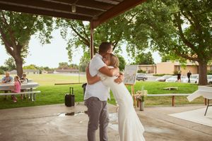 A couple dances under a rustic wooden pavilion surrounded by trees and guests at an outdoor celebration.