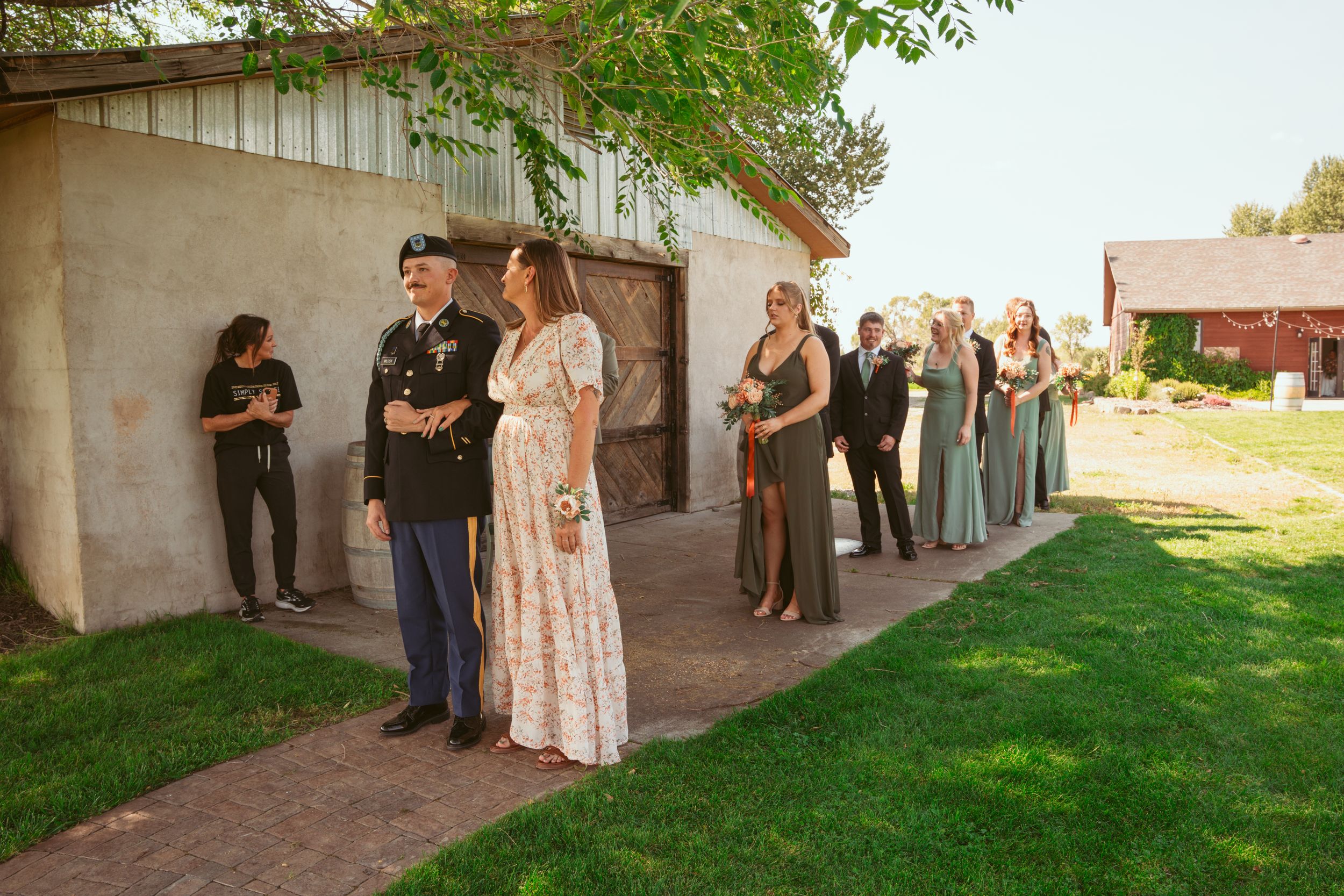 Wedding party lined up along a white concrete wall outside with green grass and trees in rural setting.