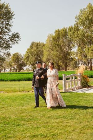 A couple walks arm in arm across a lush green lawn with trees in the background.