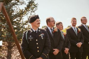 Groomsmen in formal attire stand in line outdoors against evergreen trees.