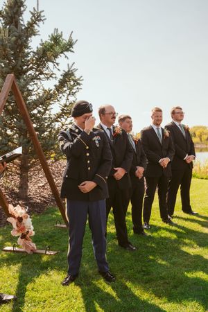 A line of groomsmen standing outdoors on grass next to evergreen trees on a sunny day.