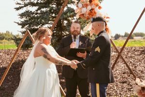 Wedding ceremony takes place under wooden arch with peach floral decorations in rustic outdoor setting.