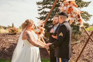 Outdoor wedding ceremony under wooden arch decorated with peach-colored flowers.