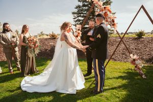 A joyful spring wedding ceremony takes place outdoors with a rustic wooden arch and pink floral decorations.