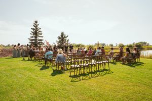 Outdoor wedding ceremony setup with wooden chairs on green lawn overlooking water under cloudy sky.