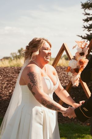 A radiant bride in a white dress laughs joyfully next to a wooden triangle arch with peach flowers.