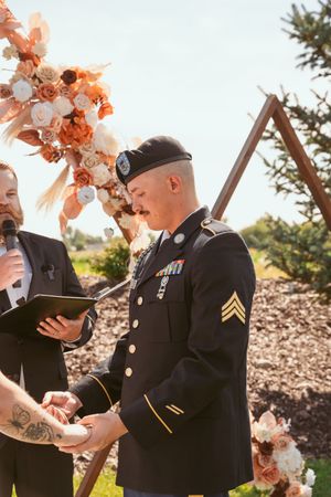 Military service members in dress uniforms stand together wearing bright coral floral headpieces during an outdoor ceremony.