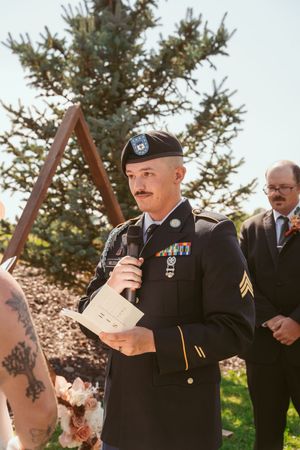 A US Army service member in dress uniform reads from papers during an outdoor ceremony.