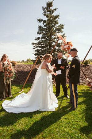Outdoor wedding ceremony under a tall evergreen tree with copper arch and pink floral decorations.