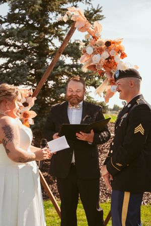 Outdoor wedding ceremony under a peach floral arch with an officiant and military servicemember in dress uniform.