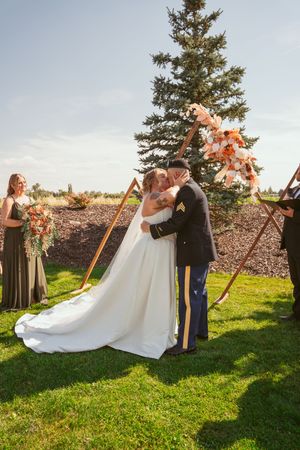 Joyful outdoor wedding ceremony beneath tall evergreen tree on sunny grassy field.