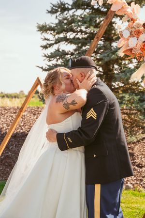 A couple share a tender kiss under a wooden archway during their outdoor military wedding ceremony.