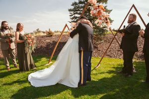 A couple embraces during their wedding ceremony beneath a wooden triangle arch on a sunny outdoor lawn.