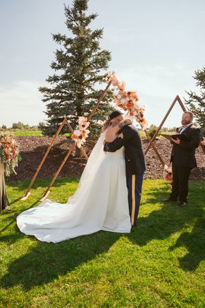 Wedding couple in elegant attire stands under floral arch decorations during outdoor ceremony at sunset.