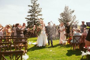 Wedding guests throw confetti as newlyweds celebrate on green lawn with pine trees in background.