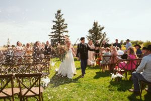 A wedding ceremony takes place outdoors with evergreen trees and wooden chairs surrounding the couple.