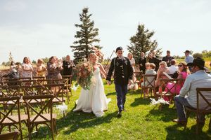 A wedding ceremony procession on a grassy lawn with guests seated in wooden chairs under pine trees.