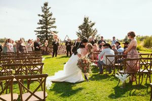 A sunlit outdoor wedding reception with rustic wooden chairs and pine trees in the background.