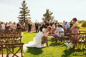 A scenic outdoor wedding ceremony on a grassy hilltop with wooden chairs and evergreen trees in the background.