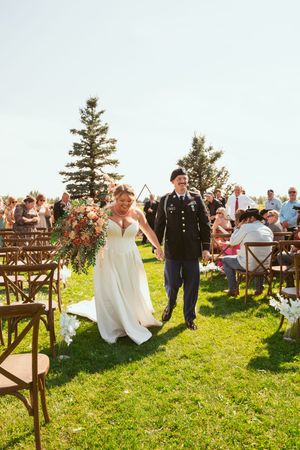 A beautiful outdoor summer wedding ceremony on a grassy hillside with wooden chairs and evergreen trees in the background.