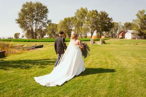 A couple in wedding attire walks across a grassy field on a sunny day with barns and trees in the distance.
