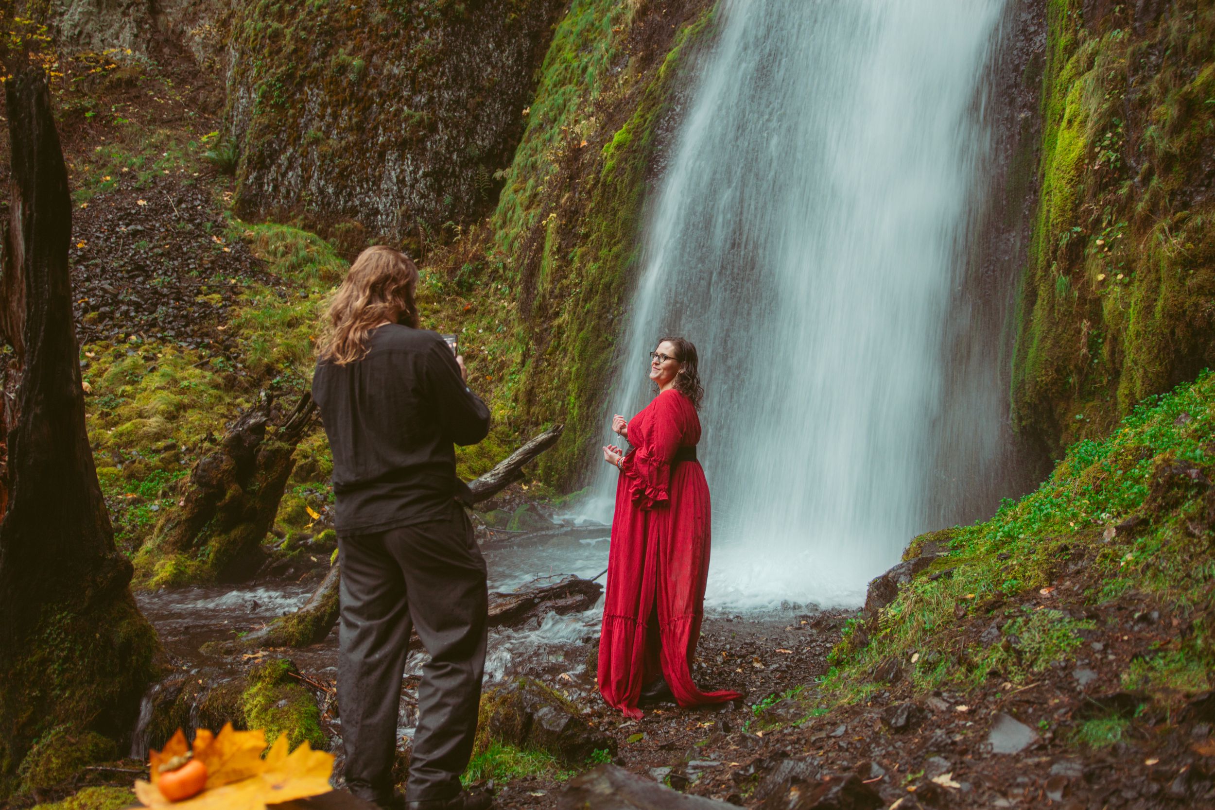 A photographer captures a scenic forest waterfall photo with a model in a flowing red dress and campfire in the foreground.