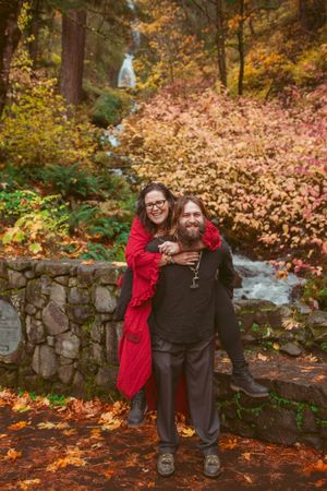 Two people embrace by a stone wall with autumn foliage and cascading waterfall in the background.