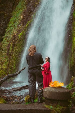 Two people stand near a majestic waterfall with moss-covered rocks and a glowing fire in the foreground.