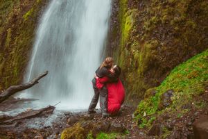 Two people embrace near a majestic waterfall surrounded by lush green moss-covered rocks in nature.