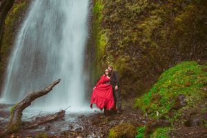 Someone in a red dress stands near a majestic waterfall cascading down moss-covered rocks in a lush forest setting.