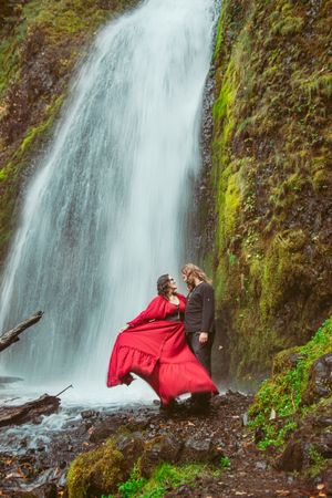 A romantic photoshoot by a cascading waterfall shows a flowing red dress billowing dramatically against the mossy rocks.