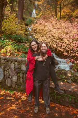 Two people embrace against autumn foliage and cascading waterfall in a tranquil forest setting.