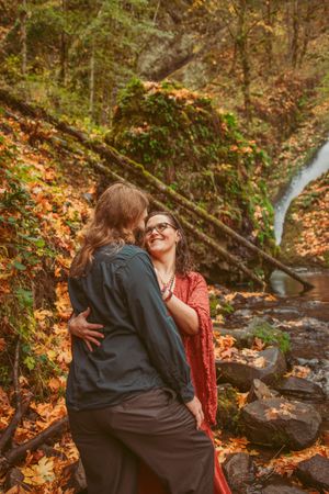 Couple embracing by a forest stream surrounded by autumn foliage and fallen leaves.