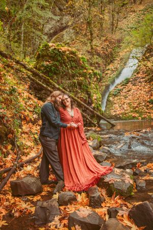A romantic autumn photoshoot by a waterfall showing a couple embracing on moss-covered rocks surrounded by fall foliage.