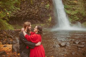 Couple embraces near majestic waterfall in scenic landscape setting.